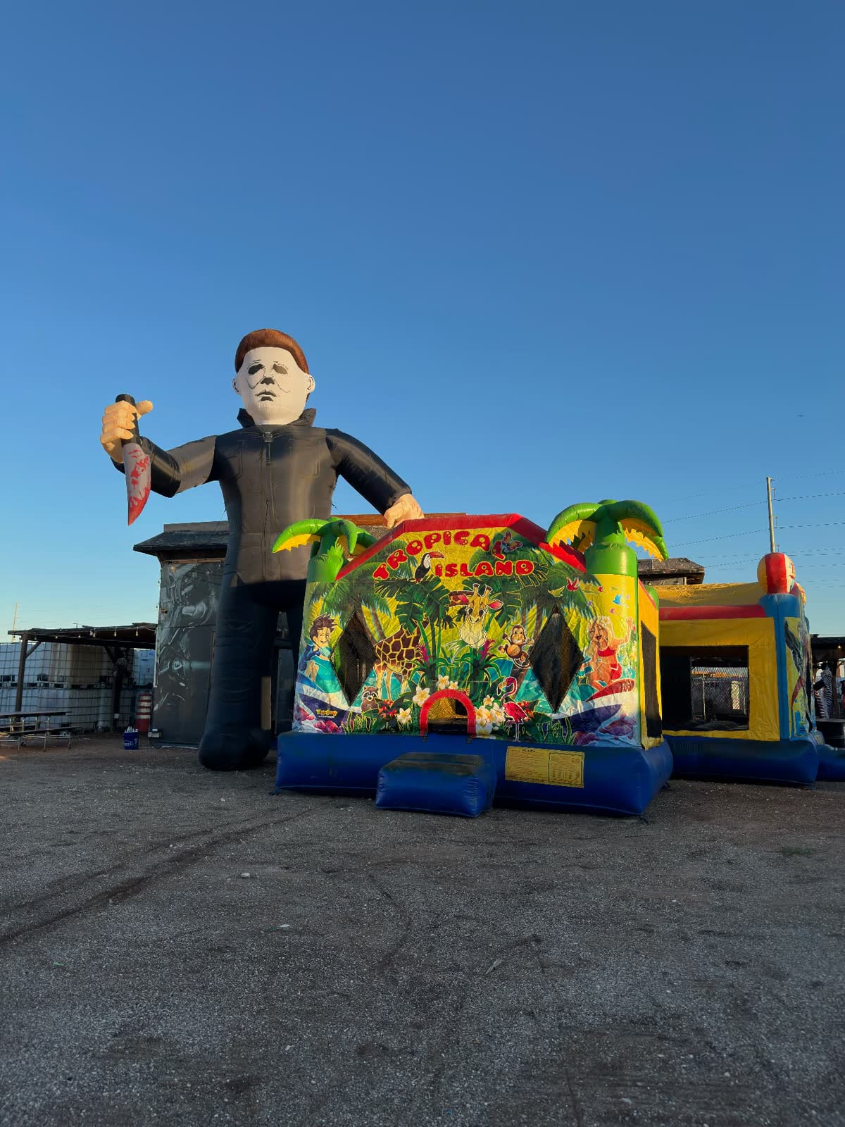 Giant masked killer inflatable towering over bounce houses on the lot before dark