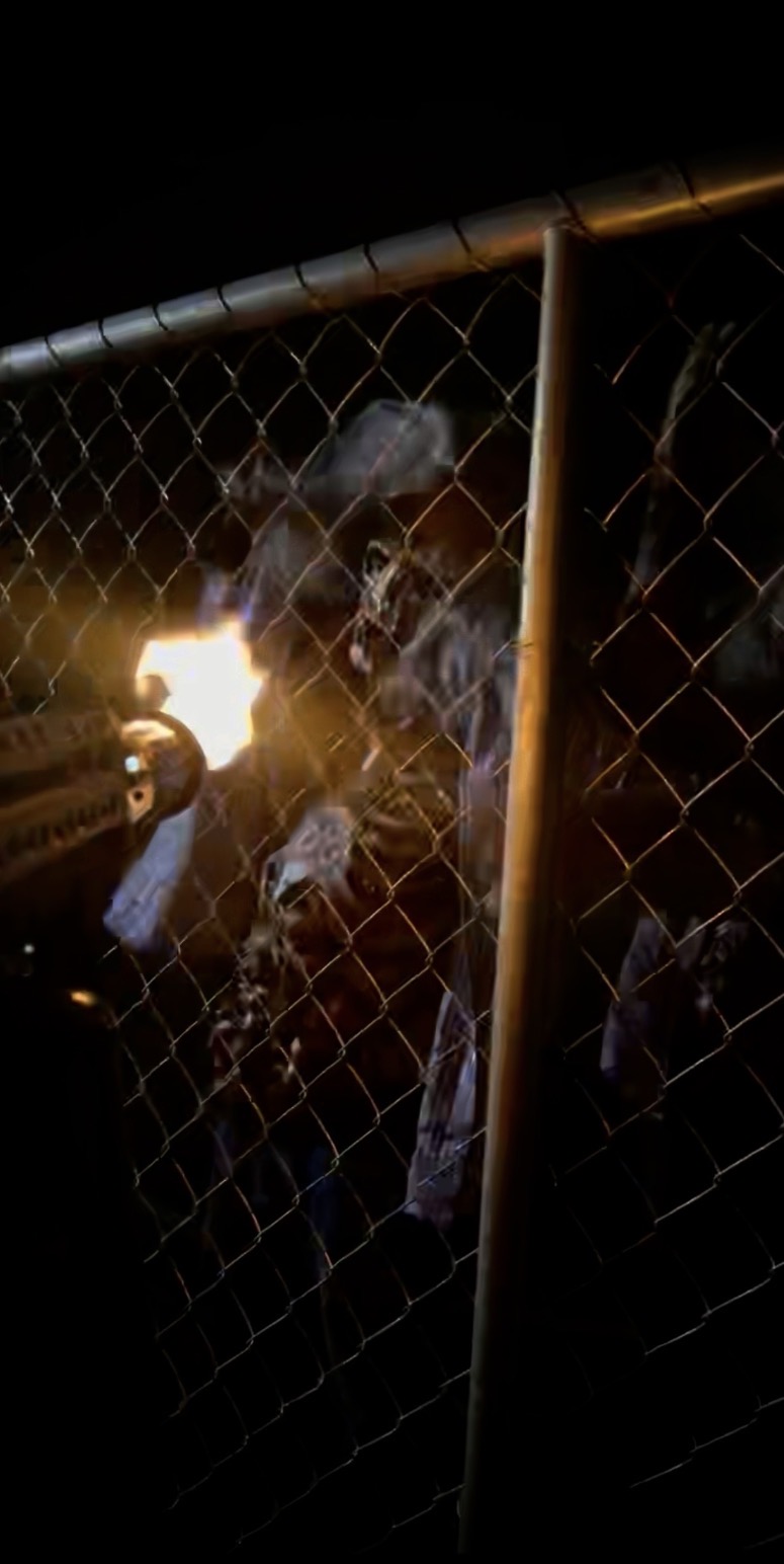 Chainsaw-wielding figure lit by sparks behind a chain-link fence